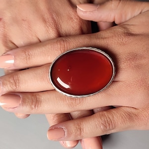 Puede incluir: Un anillo de plata con una piedra de cornalina grande de forma ovalada. La piedra es de color rojo intenso y tiene un acabado liso y pulido.