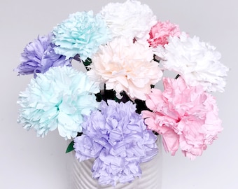 Könnte beinhalten: Ein Strauß pastellfarbener Papiercarnations in einer weißen Vase mit strukturiertem Muster. Die Carnations sind in den Farben Rosa, Blau, Lila und Weiß.
