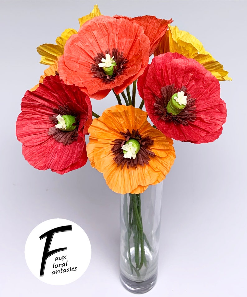 May include: A vibrant bouquet of paper poppies in a clear glass vase. The flowers are in shades of red, orange, and yellow, with brown centres and white accents. The image includes the text "Faux Floral Fantasies".