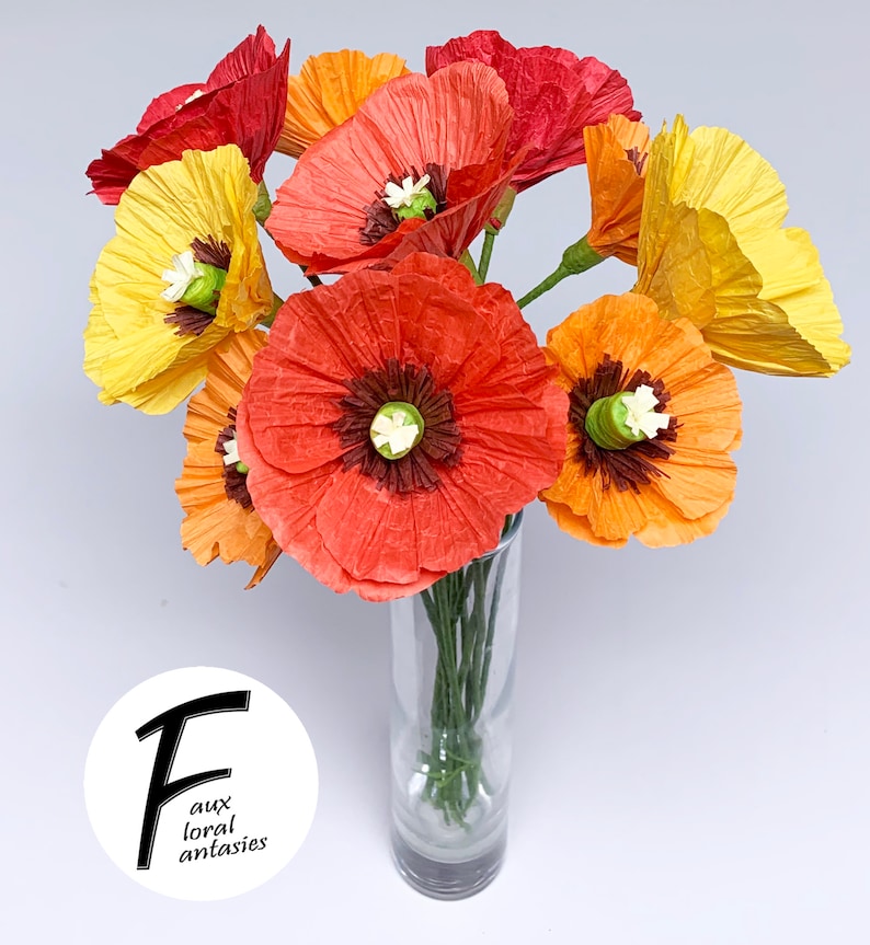 May include: A vibrant bouquet of paper poppies in a clear glass vase. The flowers are in shades of red, orange, and yellow, with green stems and white centres. The image includes the text "faux floral fantasies".