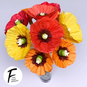 May include: A vibrant bouquet of paper poppies in red, orange, and yellow hues. The flowers feature dark brown centres and white details, displayed in a clear glass vase. The image includes the text "F aux floral fantasies".