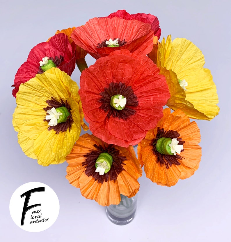 May include: A vibrant bouquet of paper poppies in red, orange, and yellow hues. The flowers feature dark brown centres and white details, displayed in a clear glass vase. The image includes the text "F aux floral fantasies".