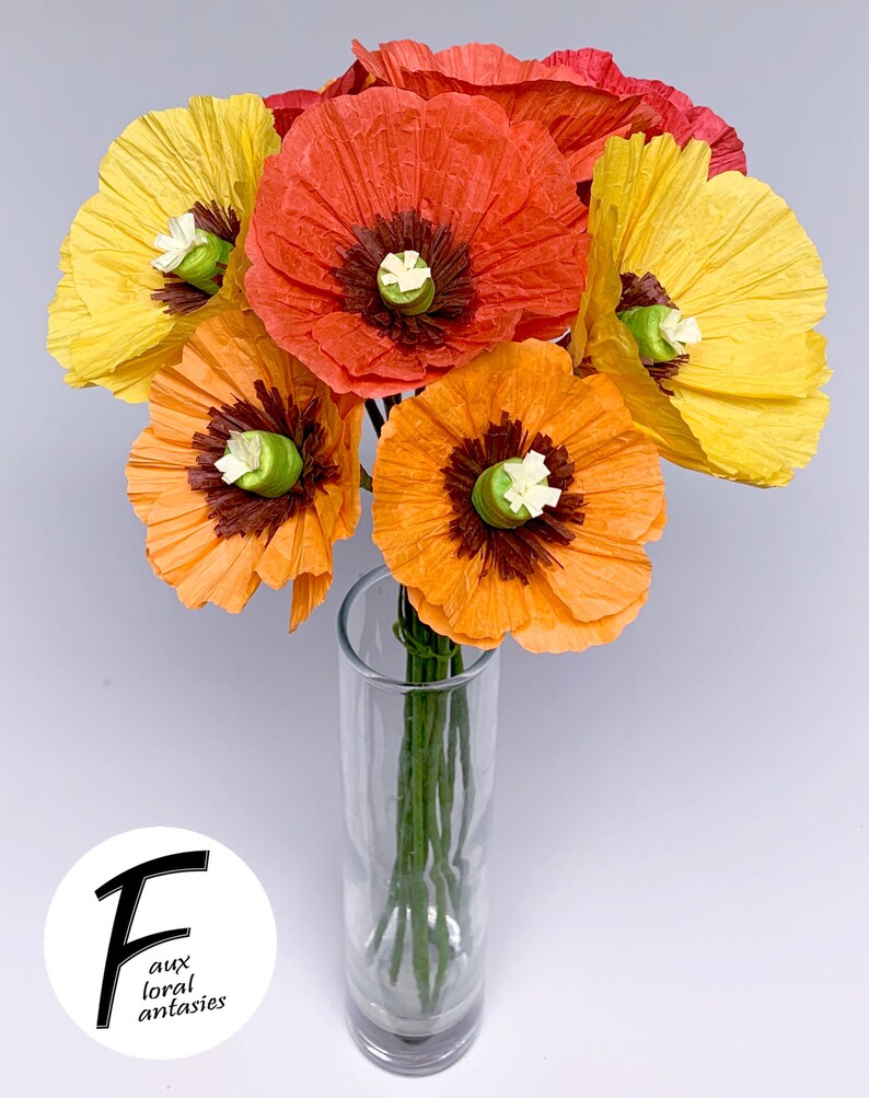 May include: A vibrant bouquet of paper poppies in a clear glass vase. The flowers are in shades of yellow, orange, and red, with dark brown centres and green stems. The image includes a logo that says "F aux floral antasies".