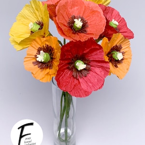 May include: A vibrant bouquet of paper poppies in a clear glass vase. The flowers are in shades of yellow, orange, and red, with dark brown centres and white accents. The vase is on a white background. The logo "Faux Floral Fantasies" is in the lower left corner.