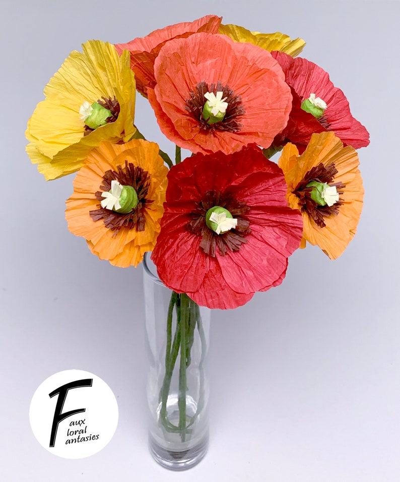 May include: A vibrant bouquet of paper poppies in a clear glass vase. The flowers are in shades of yellow, orange, and red, with dark brown centres and white accents. The vase is on a white background. The logo "Faux Floral Fantasies" is in the lower left corner.