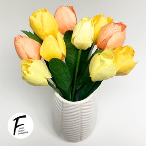 May include: A bouquet of yellow and peach paper tulips in a white vase with a textured pattern. The vase is on a white surface. The text "Faux Floral Fantasies" is on a white circle in the lower left corner of the image.