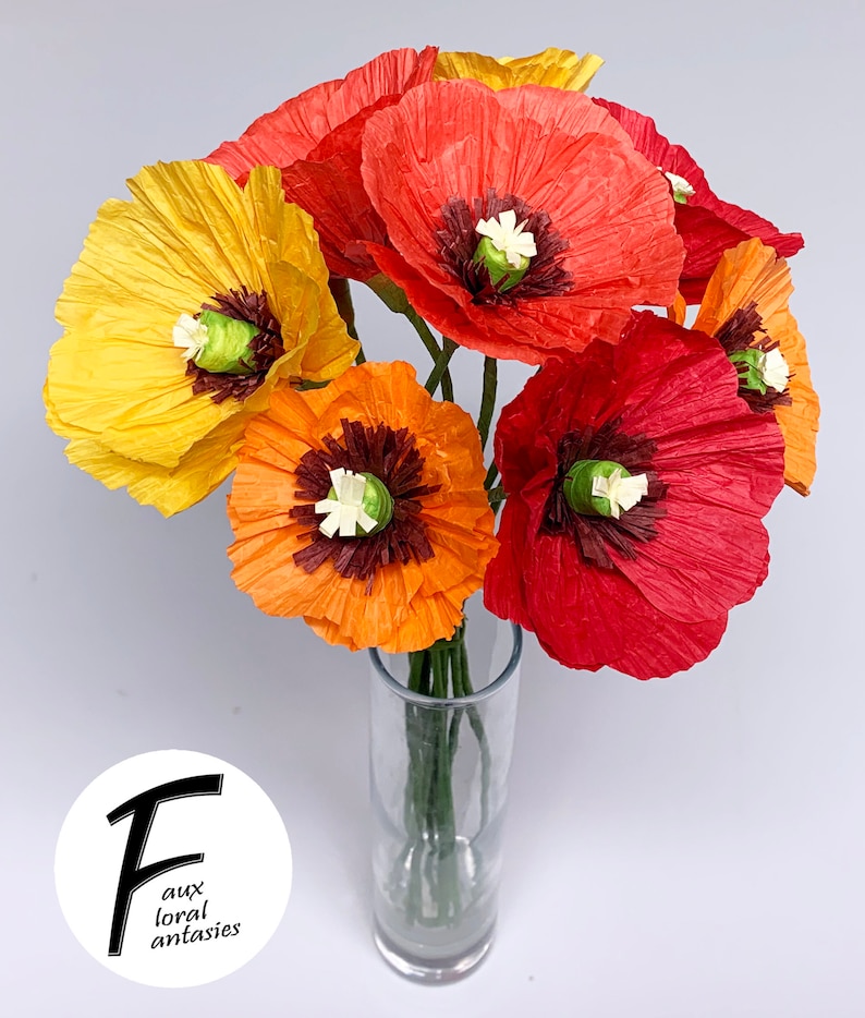 May include: A vibrant bouquet of paper poppies in a clear glass vase. The flowers are in shades of yellow, orange, and red, with dark brown centres and white accents. The vase is on a white surface. The logo for "faux floral fantasies" is in the lower left.