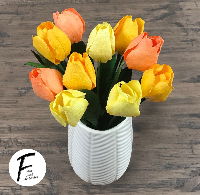 May include: A white ceramic vase with a textured design holds a bouquet of faux tulips in shades of yellow and orange.