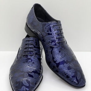 May include: A pair of navy blue dress shoes with a glossy finish. The shoes feature a detailed floral pattern and black laces. The shoes are displayed on a white surface.