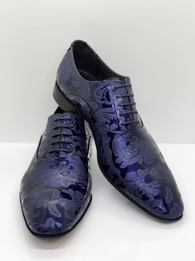 May include: A pair of navy blue dress shoes with a glossy finish. The shoes feature a detailed floral pattern and black laces. The shoes are displayed on a white surface.