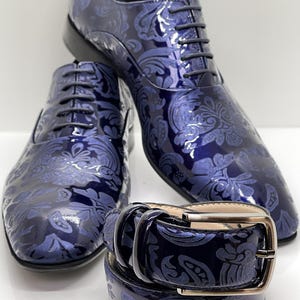 May include: A pair of navy blue patent leather dress shoes with matching belt. The shoes and belt feature a tonal floral pattern. The shoes are lace-up style. The belt has a silver-colored buckle.