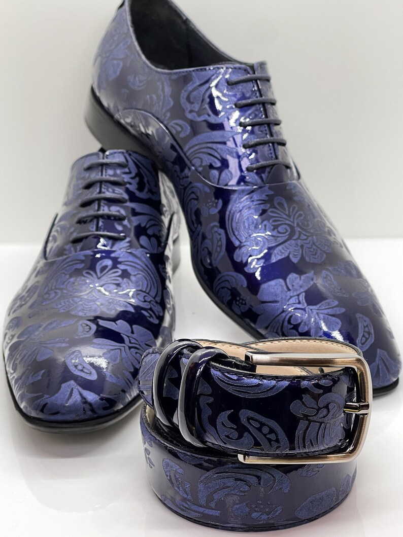 May include: A pair of navy blue patent leather dress shoes with matching belt. The shoes and belt feature a tonal floral pattern. The shoes are lace-up style. The belt has a silver-colored buckle.