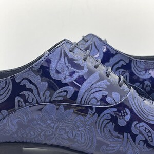 May include: Close-up of a pair of navy blue dress shoes with a glossy finish. The shoes feature a tonal, ornate floral pattern and matching laces. The shoes have a low heel and a classic Oxford style.