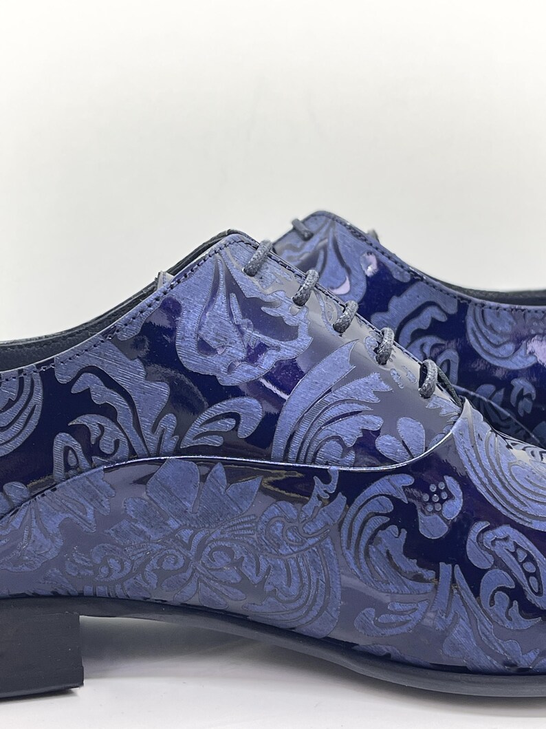 May include: Close-up of a pair of navy blue dress shoes with a glossy finish. The shoes feature a tonal, ornate floral pattern and matching laces. The shoes have a low heel and a classic Oxford style.