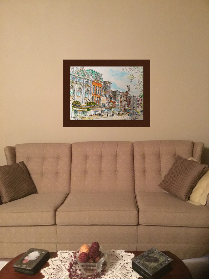 May include: A watercolour painting depicting a city street scene with buildings, trees, and people walking. The buildings are painted in shades of brown, yellow, and green. The trees are painted in shades of green and brown. The people are painted in shades of blue, green, and brown.