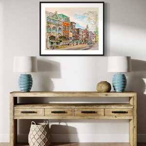 May include: A watercolour painting depicting a city street with colourful buildings. The buildings feature a range of architectural styles, including a large building with a green roof and a smaller building with a red roof. Trees line the street and there are people walking on the pavement.
