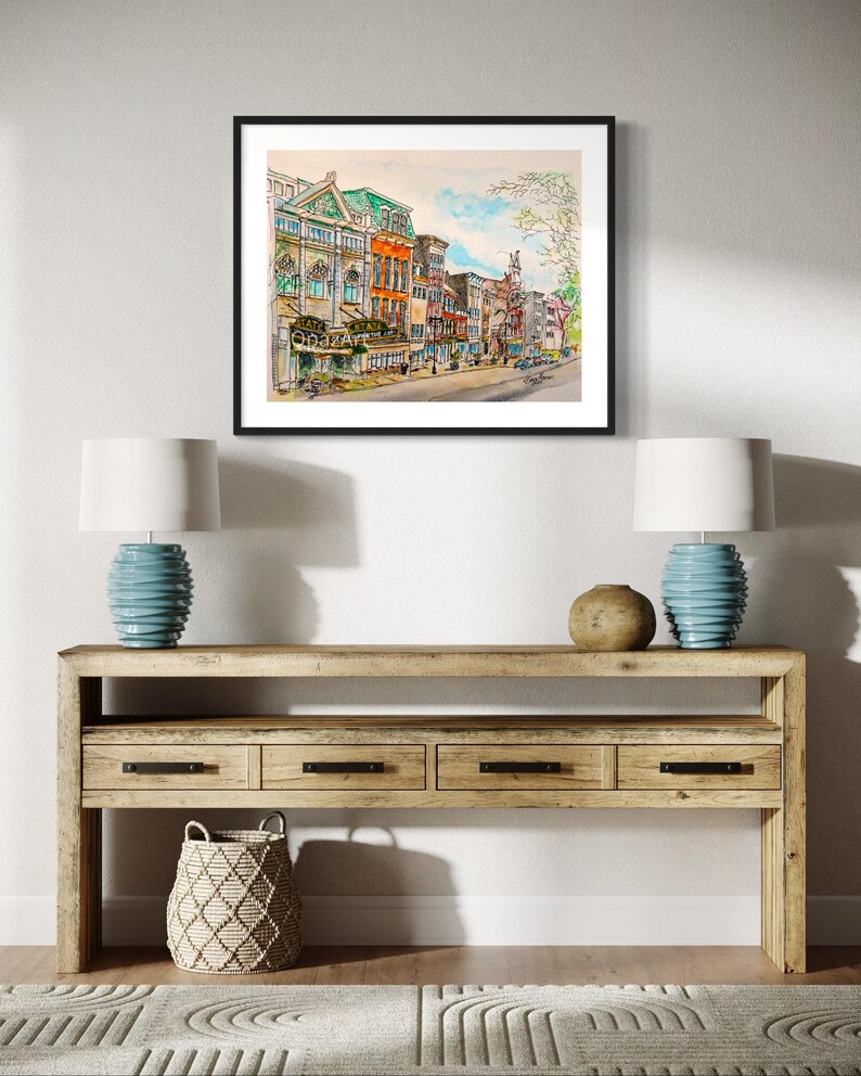 May include: A watercolour painting depicting a city street with colourful buildings. The buildings feature a range of architectural styles, including a large building with a green roof and a smaller building with a red roof. Trees line the street and there are people walking on the pavement.