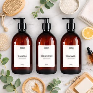 May include: Three amber plastic bottles with black pump dispensers, each featuring a white label with the text Bathroom Essentials, 500ml, and the specific product names Shampoo, Conditioner, or Body Wash printed in black.