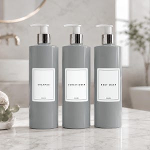 May include: Three cylindrical grey plastic bottles with white pump dispensers and silver collars. Each bottle features a rectangular white label with black text reading SHAMPOO, CONDITIONER, or BODY WASH, and the volume 500ml printed at the bottom.