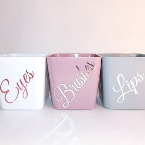 May include: Three square containers with lids, labelled "Eyes", "Brushes", and "Lips". The containers are white, pink, and grey.