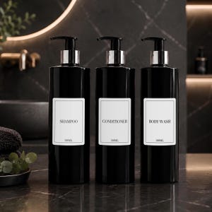 May include: Three cylindrical black plastic bottles with black pump dispensers and silver collars. Each bottle features a white rectangular label with a black border, printed with the text SHAMPOO, CONDITIONER, or BODY WASH, and the volume 500ML.