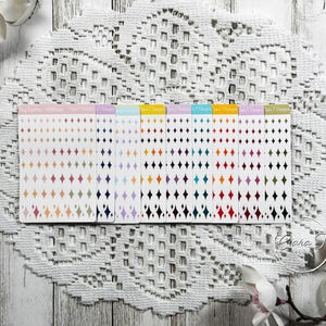 Puede incluir: Una colección de pegatinas en forma de diamante de colores dispuestas en filas sobre tarjetas blancas. Cada tarjeta presenta una combinación de colores diferente, incluyendo rosa, morado, azul y verde. Las pegatinas se muestran sobre un mantel de encaje blanco con flores blancas.
