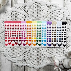 May include: A selection of heart-shaped stickers in a spectrum of colours, arranged in rows on rectangular sheets. The sheets are labelled with text and are displayed on a white, patterned surface. The stickers range from red to black.