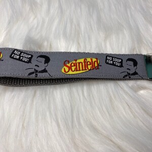 Könnte beinhalten: Ein grauer Stoffschlüsselanhänger mit einem gelben und schwarzen "Seinfeld"-Logo und einem Cartoon-Bild eines Mannes mit dem Text "No Soup for You!" darauf.