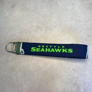 May include: A blue and white fabric keychain with a silver key ring. The keychain has the words "Seattle Seahawks" printed in green.