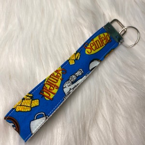 May include: A blue and yellow fabric keychain with the word "Seinfeld" printed on it. The keychain features a cartoon image of Jerry Seinfeld and a cup of coffee.