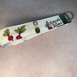 Puede incluir: Llavero blanco con una imagen de dibujos animados de Dwight Schrute de la serie The Office, con el texto "Schrute Farms" e ilustraciones de remolachas.