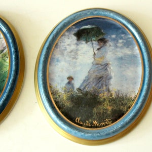 Pode incluir: Um prato de cer&acirc;mica redondo, emoldurado em azul e dourado, com uma pintura de uma mulher segurando um guarda-chuva caminhando por um campo. A pintura &eacute; assinada "Claude Monet".