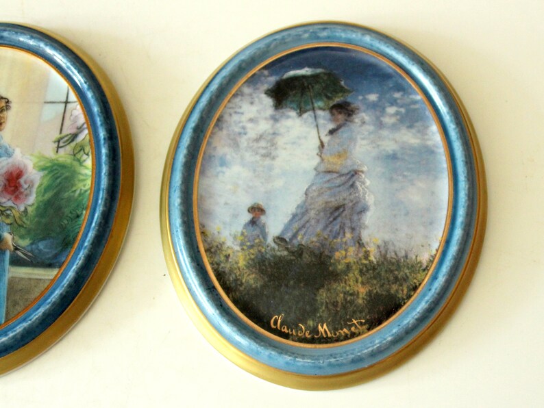 Pode incluir: Um prato de cer&acirc;mica redondo, emoldurado em azul e dourado, com uma pintura de uma mulher segurando um guarda-chuva caminhando por um campo. A pintura &eacute; assinada "Claude Monet".