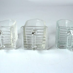 May include: Three clear glass cream pitchers with handles. The pitchers have a rectangular shape with a textured pattern on the top and sides. The handles are small and curved. The pitchers are displayed on a white surface.