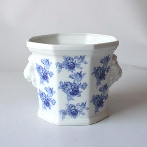 Puede incluir: Jardinera de cerámica blanca con forma hexagonal y estampado floral azul. La jardinera tiene dos asas decorativas con forma de cabeza de león. Una pieza clásica para la decoración del hogar, apta para interior o exterior.