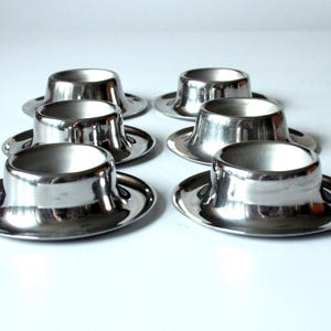 Peut inclure: Six coquetiers argentés brillants. Chaque coquetier a une base large et plate et un corps arrondi et évasé menant à une ouverture plus petite. La surface réfléchissante crée des reflets et des ombres. Disposition en deux rangées de trois.