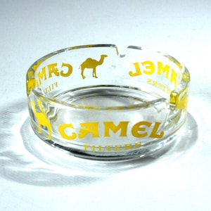 Op de afbeelding: Transparante glazen asbak met gele tekst en een kameel-logo. De asbak heeft een ronde vorm met drie inkepingen voor sigaretten. De tekst luidt "CAMEL FILTERS".