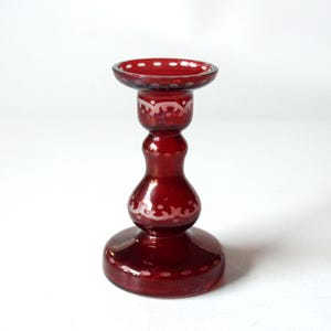 Könnte beinhalten: Ein rubinroter Glas-Kerzenhalter mit rundem Sockel, bauchiger Mitte und kleinem, kreisförmigem Oberteil. Der Kerzenhalter hat weiße Punkt- und Blumenmuster. Der Kerzenhalter ist etwa 15 cm hoch.