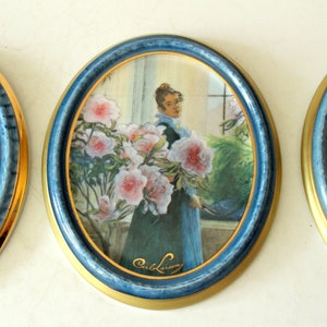 Pode incluir: Tr&ecirc;s pinturas ovais emolduradas com molduras azuis e douradas. A pintura do centro apresenta uma mulher com um vestido azul em p&eacute; em frente a uma janela com flores cor-de-rosa. A pintura &eacute; assinada "Carl Larsson".
