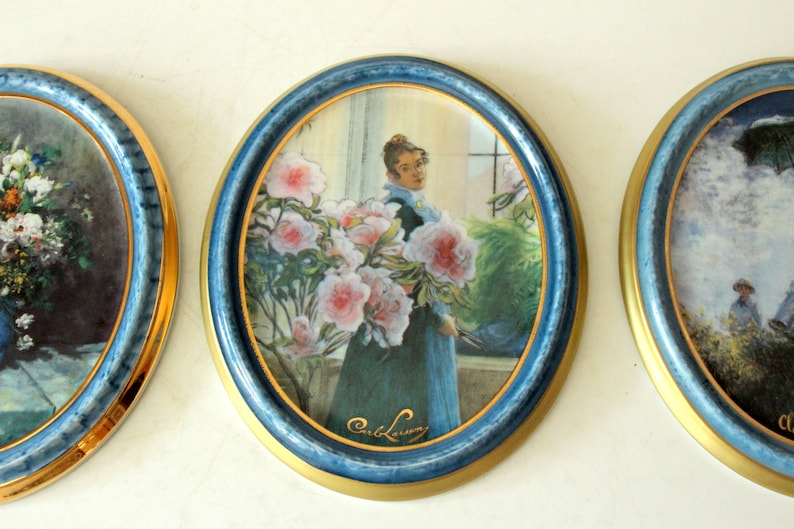 Pode incluir: Tr&ecirc;s pinturas ovais emolduradas com molduras azuis e douradas. A pintura do centro apresenta uma mulher com um vestido azul em p&eacute; em frente a uma janela com flores cor-de-rosa. A pintura &eacute; assinada "Carl Larsson".