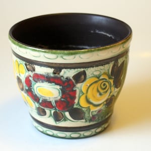 Puede incluir: Una maceta de cerámica con un borde marrón y un fondo blanco. La maceta está decorada con un patrón floral en tonos rojos, amarillos, verdes y marrones.