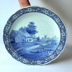 Könnte beinhalten: Ein großer blauer und weißer Keramikteller mit einer holländischen Landschaftsszene. Der Teller zeigt ein Bauernhaus, Windmühlen, eine Brücke und einen Fluss. Der Rand des Tellers ist mit einem floralen Muster verziert.