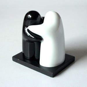 Könnte beinhalten: Ein schwarzes und weißes Keramik-Salz- und Pfefferstreuer-Set. Die Streuer sind wie sich umarmende Figuren geformt, eine schwarz und eine weiß, auf einem schwarzen rechteckigen Sockel montiert. Das Design ist minimalistisch und modern.
