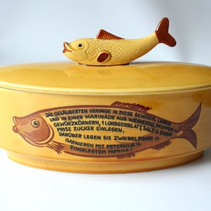 Peut inclure: Plat de service en céramique jaune avec une poignée en forme de poisson sur le couvercle. Le plat présente une illustration de poisson marron et du texte en allemand. Il a deux poignées.
