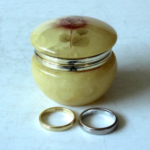 May include: A small, round, yellow alabaster ring box with a floral design on the lid. Two wedding bands are in front of the box, one gold and one silver.