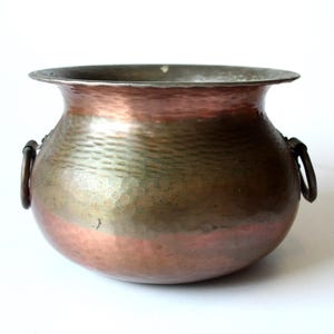 Könnte beinhalten: Ein gehämmertes Kupfergefäß mit breitem Rand und zwei Eisengriffen. Das Gefäß hat einen abgerundeten Körper mit einem rotbraunen Boden, der in eine grün-bronze Farbe übergeht. Der Rand ist ein dunkleres, angelaufenes Metall.