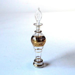 May include: A clear glass perfume bottle with a gold design. The bottle has a bulbous center with gold detailing and a clear, elongated stopper. The base and neck are also accented with gold bands.