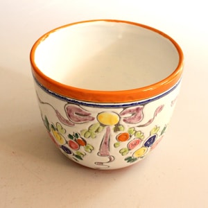 Könnte beinhalten: Eine weiße Keramikschale mit orangefarbenem Rand und floralem Design. Die Schale hat ein dekoratives Muster aus Blumen, Bändern und einem gelben Zentrum. Der Rand hat einen blauen Streifen unter dem orangefarbenen Rand.