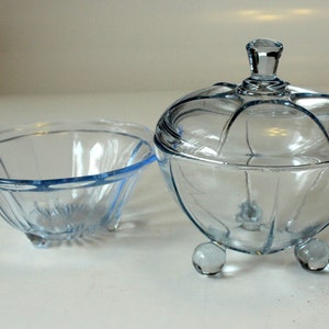 May include: A set of two clear glass bowls with a light blue tint. The larger bowl has a lid and sits on three feet. The smaller bowl has a scalloped edge and sits on three feet.