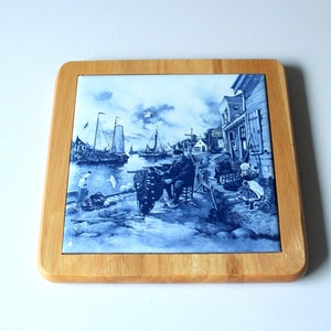 Può includere: Piastrella quadrata in ceramica con una scena nautica blu e bianca, incorniciata in una cornice di legno chiaro. L'immagine raffigura un porto con barche a vela, edifici e persone. Un pezzo decorativo per la casa.